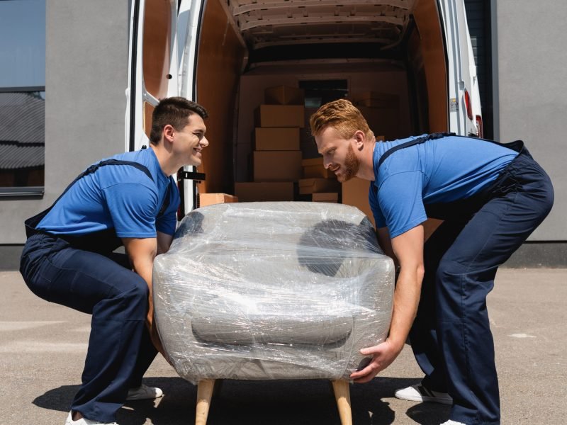 Side view of movers holding armchair in stretch wrap near track on urban street